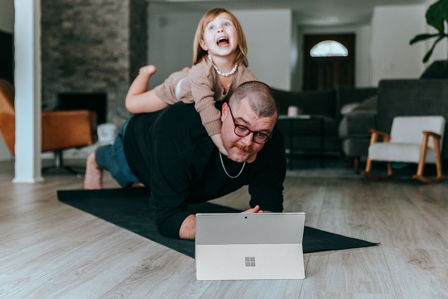 A man tries to hold a plank in front of a laptop while his child hangs onto his neck.